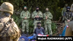 Польскія (на пярэднім пляне) і беларускія (на заднім пляне) памежнікі стаяць побач з мігрантамі, якія, як мяркуецца, паходзяць з Афганістана і сядзяць на зямлі ў невялікай вёсцы Уснаж-Гурны каля Беластока, на паўночным усходзе Польшчы, разьмешчанай недалёка ад мяжы. 2021